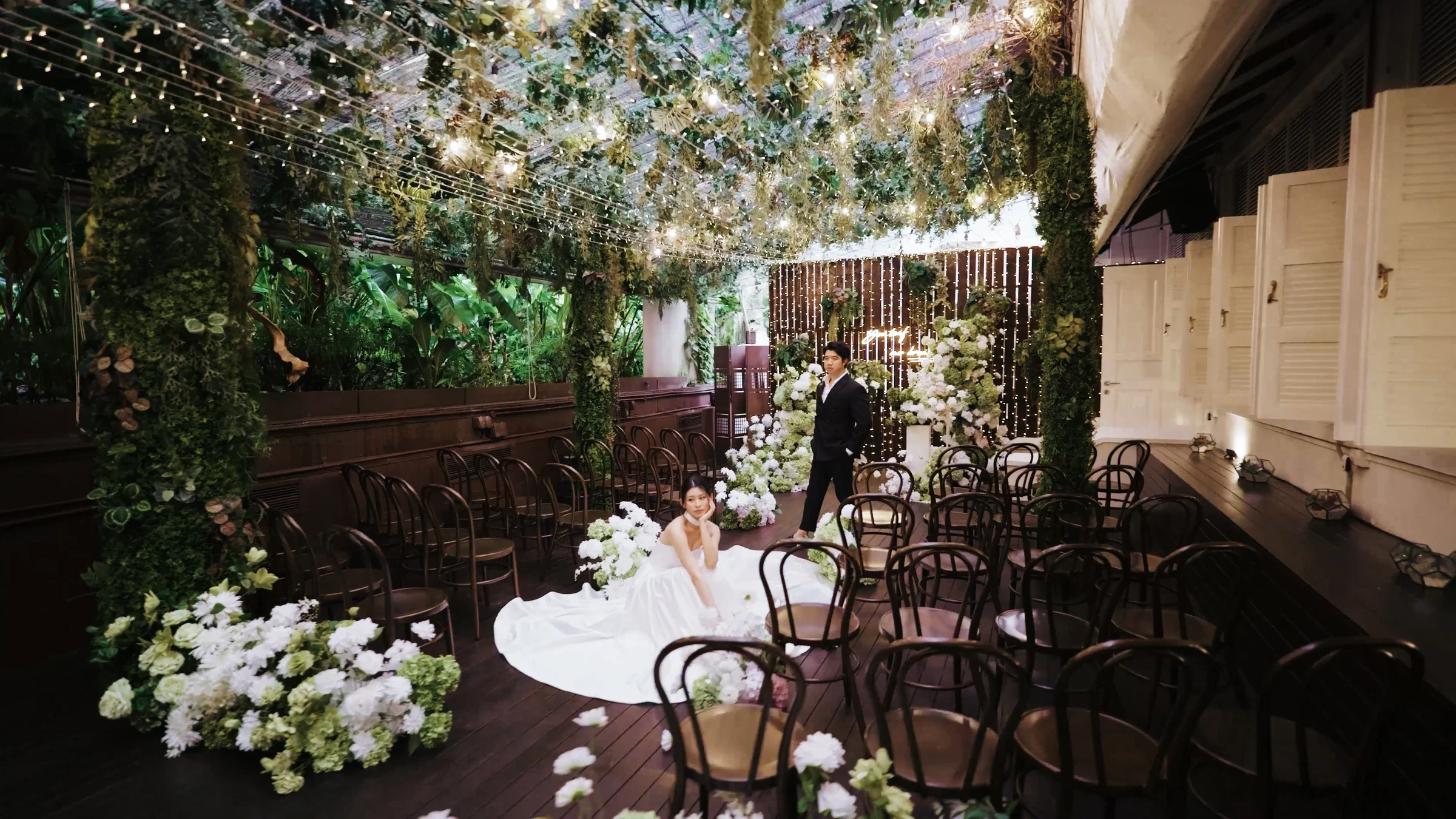 Bride and groom surrounded by flowers, greenery, and hanging lights at a wedding venue